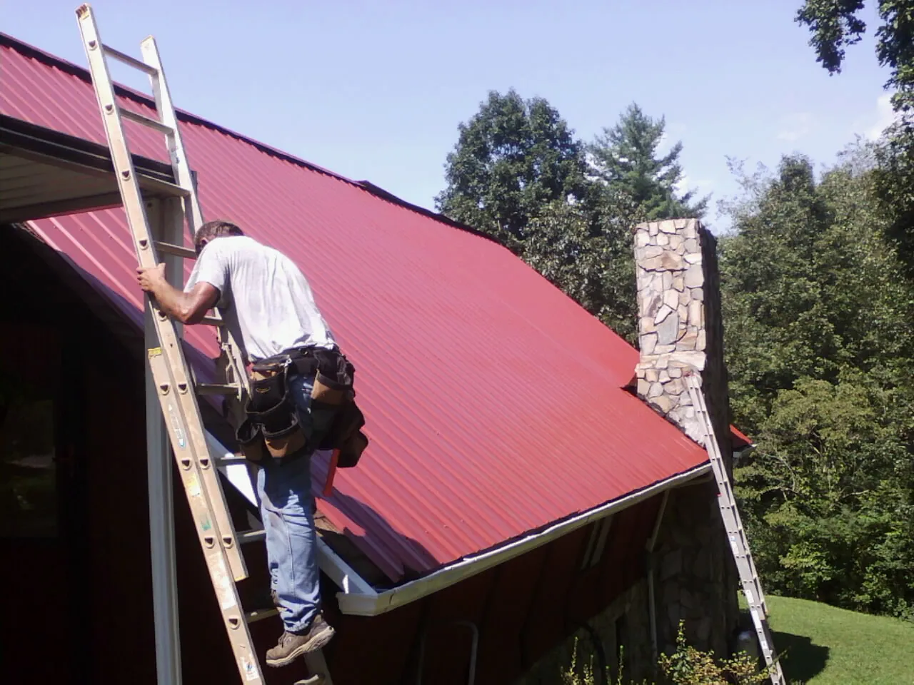 Roofer on a ladder beside a red metal roof and stone chimney — Soffit & Fascia Repair by Roofing Brothers in Steen
